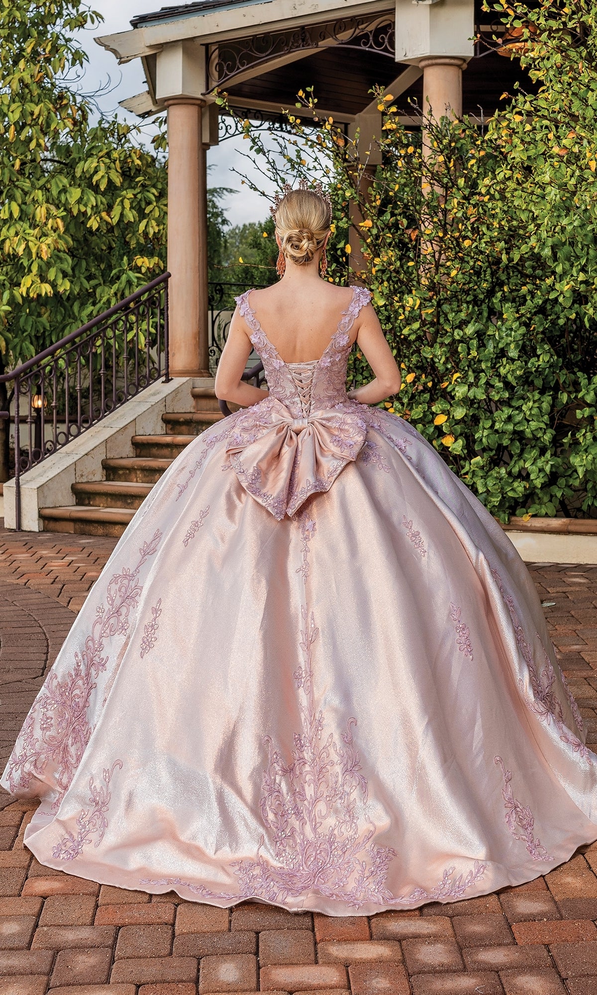 A woman stands outdoors facing away, wearing the Quinceanera Dress 1762 By Dancing Queen—a pink ball gown with intricate embroidery, a large back bow, and an elegant a-line silhouette—by steps and greenery.